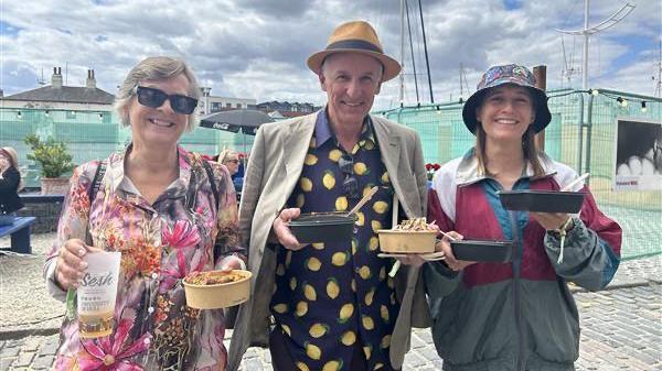 Three people stand on a cobbled street holding food cartons. From left to right: A woman with short light-coloured hair wearing a multi-coloured patterned blouse and black sunglasses; a man wearing a beige sunhat, a beige blazer draped over his shoulders and a navy shirt with lemons on; a woman with shoulder-length brown hair wearing a black patterned bucket hat and a burgundy, blue ad grey jacket. 