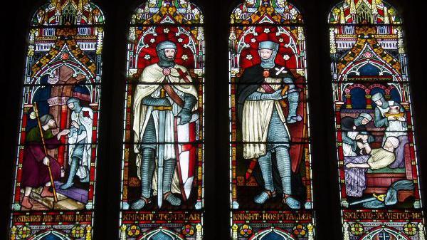 Stained glass windows. The four windows feature knights. Two in the centre show individual knights. One on the left shows a knight speaking to a man who holds a cane. The knight is holding the man's hand and is standing above him on a step. In the window on the right two knights are caring for a man who is lying in bed. One knight is holding a jug of water.