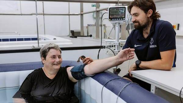 A smiling woman sitting in a hot tub in what appears to be a research lab with white walls. A man standing next to the tub is taking her blood pressure