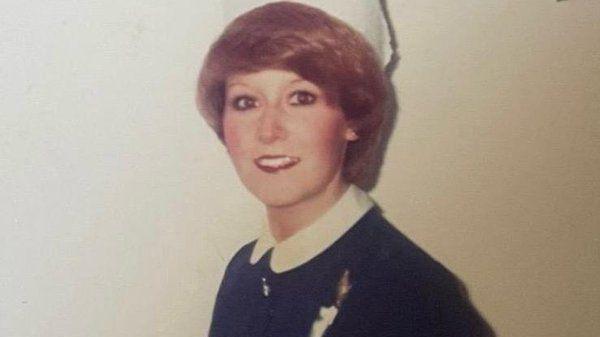 The Image shows a women in a 1970s nurse work outfit, she has big orange hair and is wearing lots of black eye makeup.