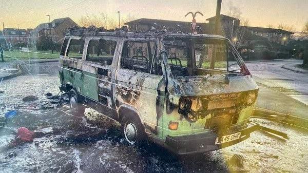 The Campervan after the fire. It is green and white and parked in a car park. It has suffered severe fire damage. Paint has peeled away, its windows have been smashed and its lights destroyed. Its tyres are flat. There is burnt debris on the ground around it.