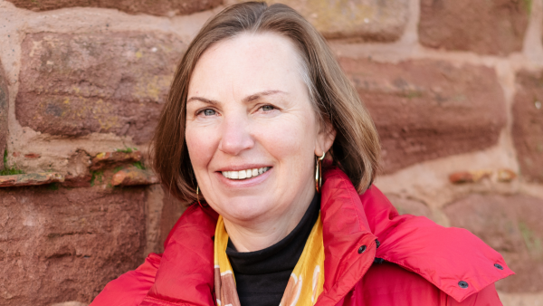 A woman with shoulder length fair hair is standing by a sandstone wall. She is wearing a red coat, black top, yellow scarf and gold hoop earrings and smiling at the camera.