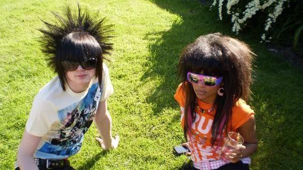 A boy on the left wears a white T-Shirt with blue designs on the front. He wears large brown sunglasses and has a straight black fringe. The back of his head is styled to show spikes. A woman on the right has an orange T-Shirt with writing on and carries a cup. She wears large reflective pink and purple sunglasses. Her hair is brown and large, she has a straight fringe and pink hair extensions. 