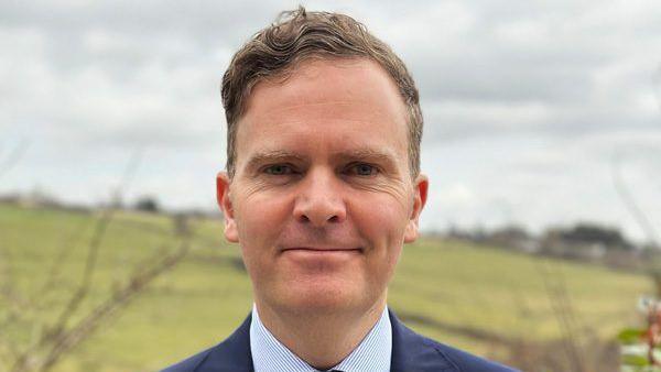 Ross Lambie, with short fair hair swept to one side, looking directly at the camera. He is smiling. He is wearing a blue suit over a blue and white striped shirt. Only his shoulders and collar are visible. He is standing in front of a blurred background with a green hill.