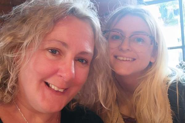 A mother taking a selfie with her adult daughter. The mum has curly blonde hair and is smiling on the left of the frame. Her daughter, also smiling, has long blonde hair and wears wide-rimmed glasses. They are standing in front of a window with light streaming in.
