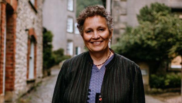 A woman with short curly hair wearing a blue top and green jacket stands in a cobbled street.