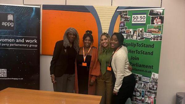 A group of women including Mrs Soanes stand at the end of a room near several poster boards and smile at the camera. A table is in front of them.