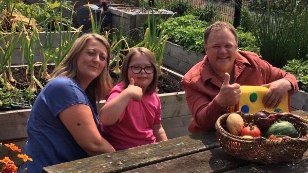 Three people sitting on a bench in a garden. Left to right: Assistant head teacher Clare Charlesworth, pupil Ellie, and TV presenter Justin Fletcher