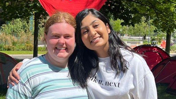 Two women are smiling at the camera with their arms around each other. The woman on the left has tied back red hair and is wearing a green and blue stripped top. The woman on the left has long dark hair and is wearing a white sweater with the words 'Thank u, next' on the front. They are standing in front of tents with some bushes and grass in the background.