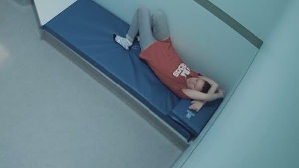 Still from CCTV footage that shows Sacha Goulden in a red Adidas T-shirt and grey leggings laying down on blue mattress in a custody cell. She has her hands folded above her head and her knees in the air.