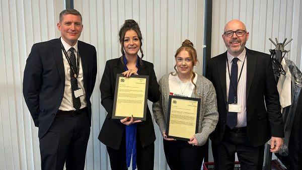 Two women smile and hold up framed certificates. They are flanked either side by men in suits.