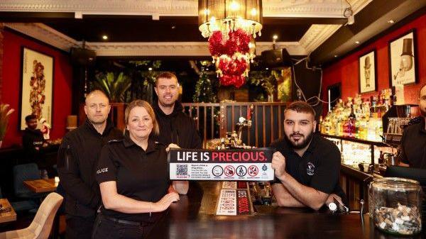 Staff at The Cherry Tree pub in Darlington. Two members of staff are holding one of the bar runners which contains the QR code. The runner reads, life is precious.