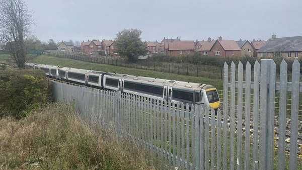 The train travelling on the route. In front is a silver metal fence, and behind it is a housing estate.