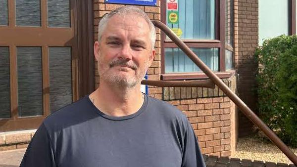 Alan Dobson standing on the steps outside the We Are With You offices. He has cropped grey hair and a short stubble beard and is wearing a black t-shirt