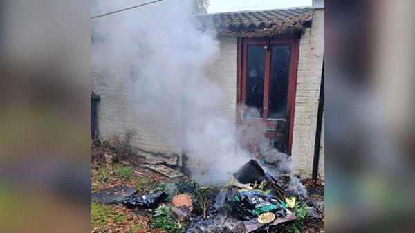 A pile of rubbish on a grassy area in front of a wooden door in a brick wall, with smoke rising from the pile