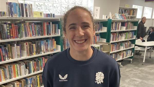 Abby Dow smiling at the camera in a navy England rugby shirt. She is standing in a library.