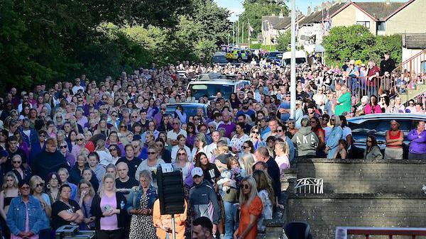 A large group of people, many wearing purple, stand in the street outside Sarah Montgomery's house.