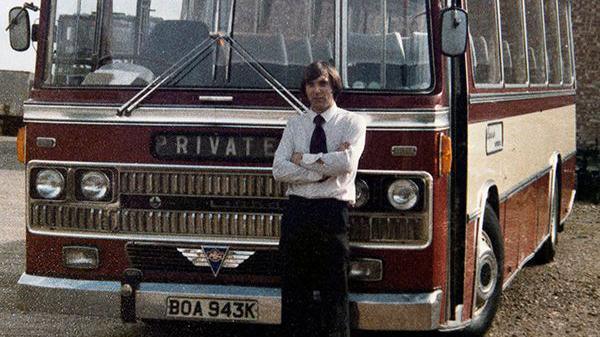 Roger Gretton standing in front of a coach, wearing a shirt and black tie