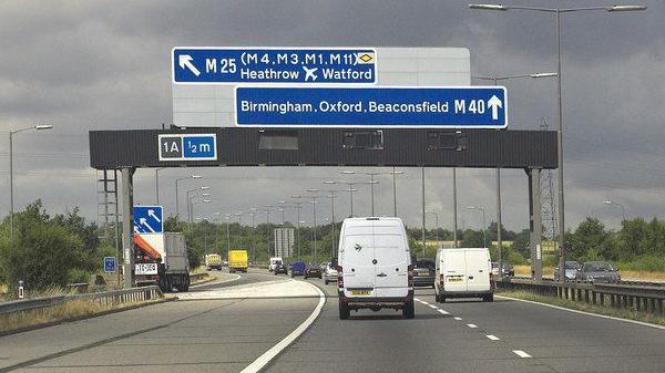 Vehicles in two lanes on the M40 travel under a large blue sign that directs them either to Heathrow and Watford or Birmingham, Oxford and Beaconsfield. The picture has been taken on a stormy day.