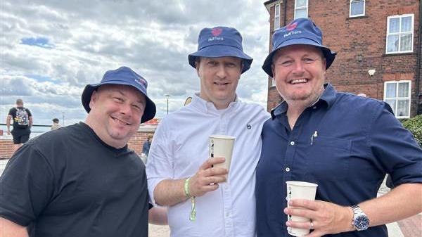 Three men wearing navy bucket hats with a Hull Trains logo on. The man on the left is wearing a black t-shirt, the man in the middle is wearing a white shirt and is holding a cup, the man on the left is wearing a navy shirt and is also holding a cup.