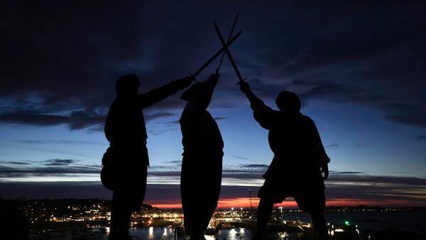 Three silhouettes of men holding swords above their heads at dusk with the lights of Guernsey in the background