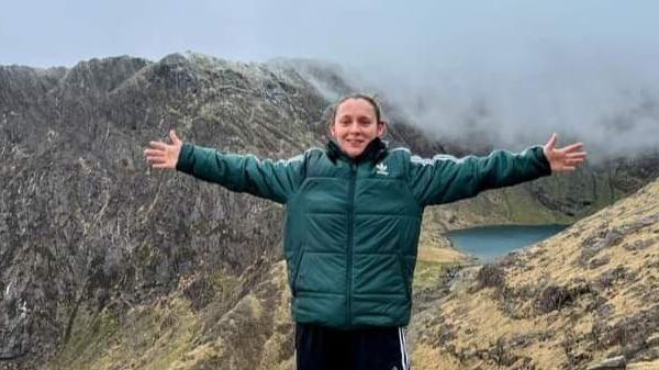 Elissa Hubbard in a green sports jacket and black Adidas tracksuit bottoms stands at the top of a mountain and a lake is also visible in the background. She has her arms outstretched and is smiling 