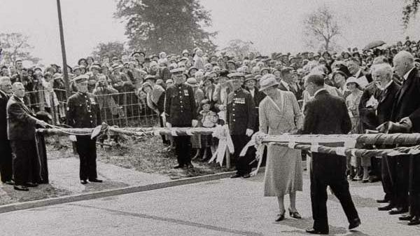 A black and white photo of a woman in light dress and hat, cutting a ribbon on road barriers