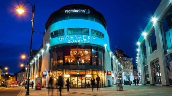 The Brewery Quarter in Cheltenham lit up at night. The main building shown in the picture is rounded at the corner facing the camera, and shows a cafe on the bottom floor, with a Cosy Club and a Premier Inn above it. People are walking by.