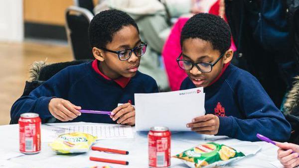 Two young male students, wearing glasses and blue school uniforms, work on some tasks. The table they are working on includes drinks, snacks and stationery. 