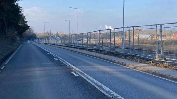 The A134 with one car on it in the very far distance, but a long row of metal fencing, showing a housing estate being built. There are two lanes of the road, and a large blue sky, with street lights down one side. 