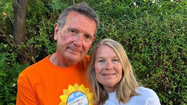 Haydn and Jayne Morgan are pictured wearing charity t-shirts with their logo which is a sunflower with Bronwen's Wish written on it. Hayden, who has an orange t-shirt, has his arm around Jayne, who is wearing a white t-shirt, and they are standing in front of a hedge.