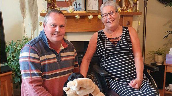 Ian Bell crouches next to his wife as they smile at the camera. They're black dog sits in the middle of them holding a teddy in its mouth. Mr Bell has short dark grey hair and wears a blue, white and red striped jumper. Mrs Bell wears and navy and white striped dress.