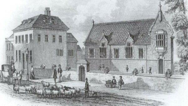 A black and white historical image of the building. The date is not known but there are sheep in the road and a horse and carriage, and men are walking on the pavement with top hats and canes with boys from the school nearby.