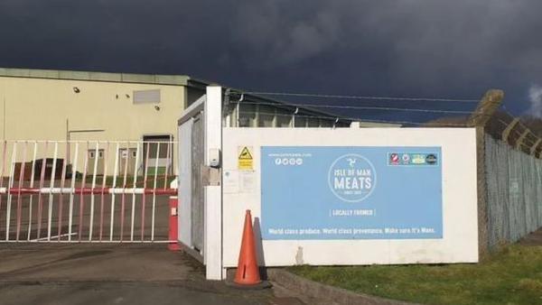 The Isle of Man Meat Plant, which is a cream coloured flat roofed building. It's surrounded by fencing and a red and white metal gate, with a large blue rectangular sign with the Isle of Man Meats logo on it on white.