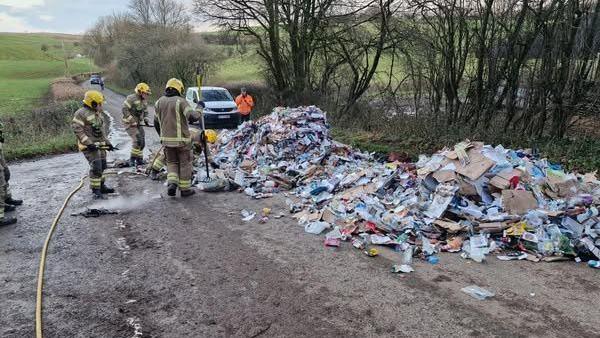 Three fire officers  stand next to two large piles of rubbish on the road. One of them is pointing a fire house at one of the piles which has been recently extinguished