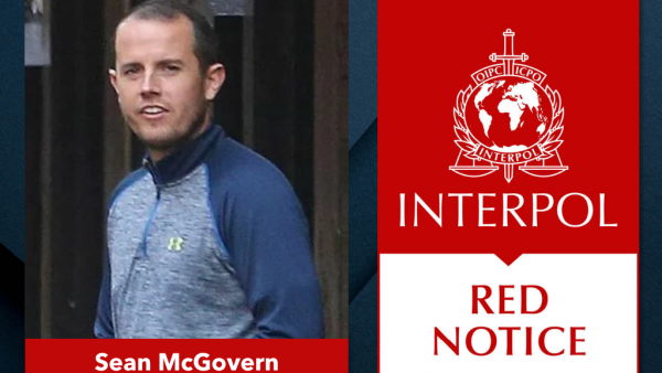 A composite image showing a picture of Sean McGovern - a man with short brown hair wearing a blue and grey zip up hoodie - on the left, on the right is the Interpol logo in white against a red background with Red Notice written underneath in a red font against a white background. His name 9Sean McGovern' is written in white with a red background underneath him