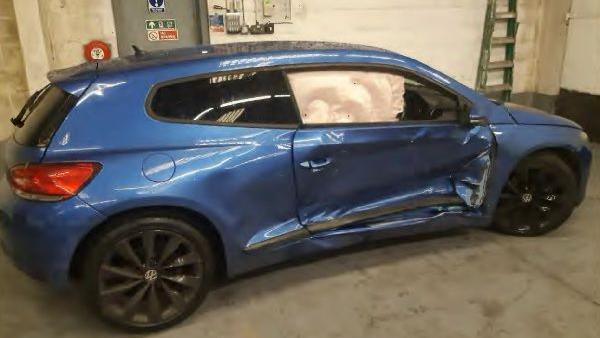 A blue hatchback bar in a garage with extensive damage to the driver's side doors, where the panels have been crushed inwards