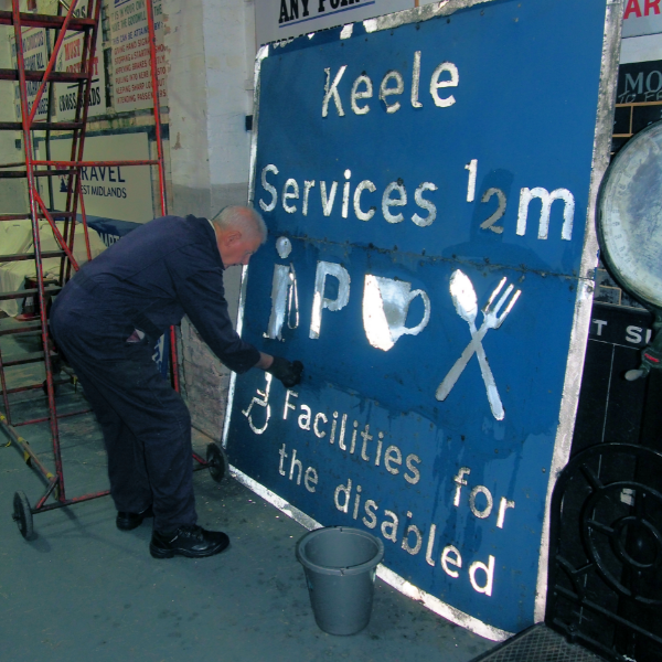 The Keele Services road sign, which is blue with white writing and a white border, which is being painted on by a man with silver hair and a blue boiler suit, who is bending down to paint it