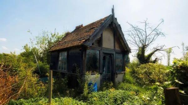 A dilapidated looking building sits amid thick vegetation. It has a panelled window and door and a tiled roof.