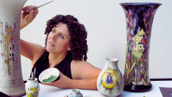 A woman can be seen against a white backdrop, painting tall pieces of pottery. She is wearing a black sleeveless top and is holding a bowl of green paint.