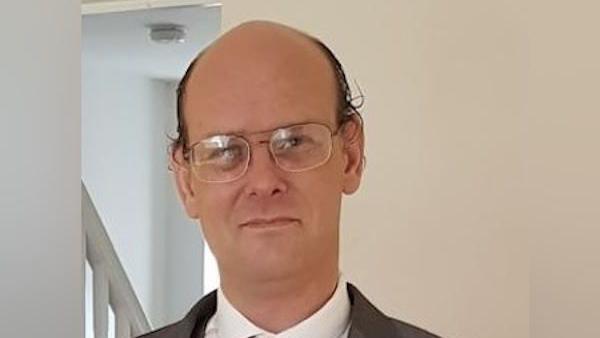 A man with a bald head and large, wire-framed glasses looks at the camera. There is a light reflected in the lenses of his flasses. He has brown, slightly curly hair at the sides of his head. He is wearing a white collared shirt and a dark jacket, which is not fully visible at the bottom of the photo.