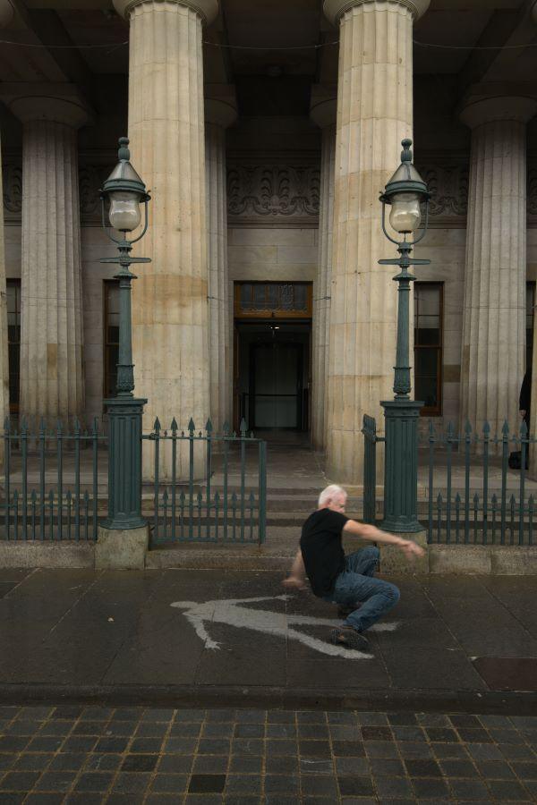Andy Goldsworthy is got in mid action from getting up from the wet ground. There is an imprint of his body shame on the pavement that is dry.