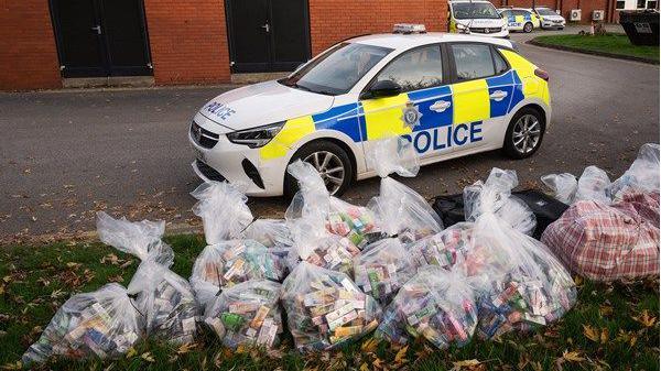 A photo of multiple clear plastic bags of illegal vapes and cigarettes on the grass in front of a parked police car. 