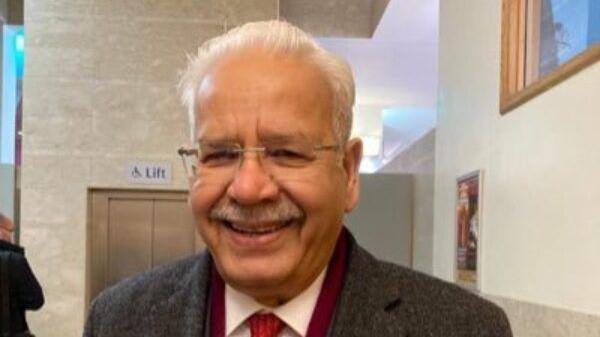 A man smiling with white hair and a moustache. He is in the lobby of a building and wearing a grey coat.