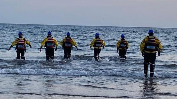 Six coastguard officers can be seen in full uniform running into the sea. They all wear life jackets and yellow jackets with blue helmets.