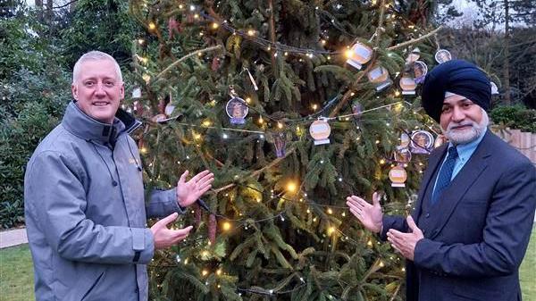 Two men standing in front of a Christmas tree with fairy lights and paper baubles. The tree is outside, on a grassy lawn. Both men are gesticulating towards the tree. The man on the left has grey hair and is wearing a grey jacket. The man on the right is in a three-piece navy suit, royal blue tie, blue shirt and is wearing a black turban. He has a grey beard.