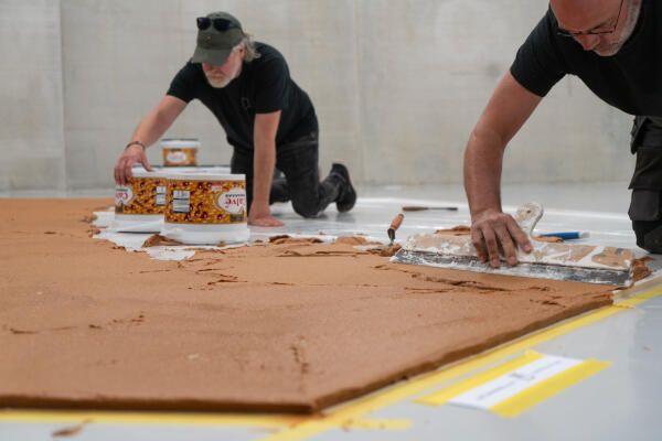 Wim T. Schippers, Peanut Butter Floor, 1962. Making process in 2026 at the Depot of Museum Boijmans Van Beuningen. © Raaf Blanker. Two men are seen spreading peanut butter on the floor of a gallery.