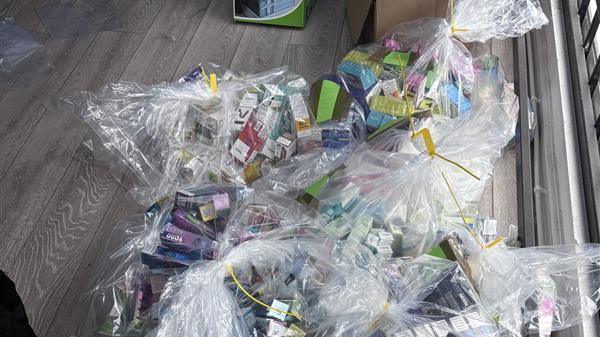 Clear plastic bags filled with colourful items, many in small cardboard boxes. They appear to be packets of vapes and cigarettes. They are coloured purple, green, blue and yellow. Yellow cable ties bind the plastic bags, which are sitting on a laminate wood floor.