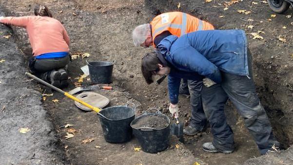 Four people are working in a shallow muddy trench in the ground. They are working with small and large shovels. Buckets of dirt are around them.