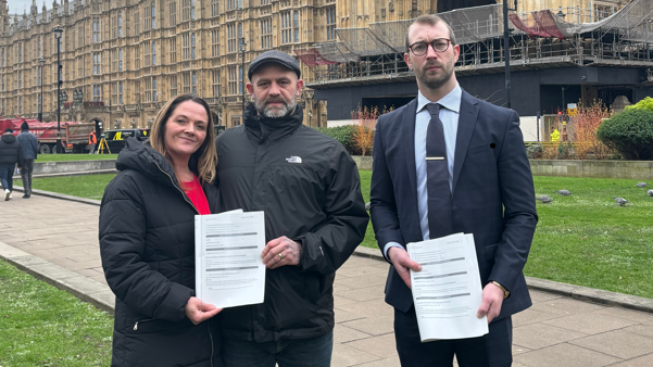 Three people standing in front of the House of Commons, holding two copies of a bill 